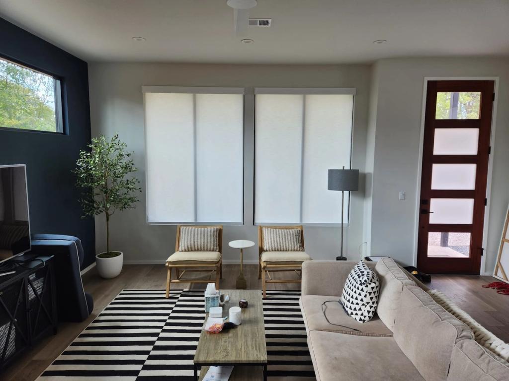 White roller shades on two large windows in a modern living room with neutral furnishings