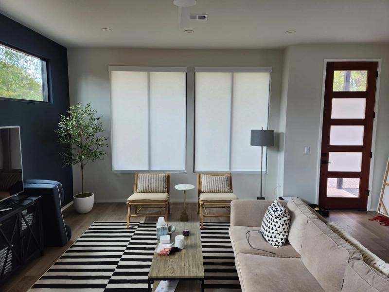 White roller shades on two large windows in a modern living room with neutral furnishings