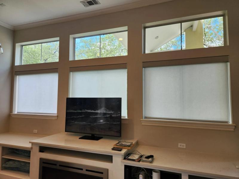 White roller shades partially lowered on three tall windows above a built-in media console with TV