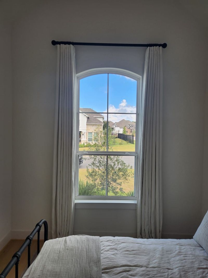 Ivory pinch-pleat drapes on black rod framing arched bedroom window with neighborhood view
