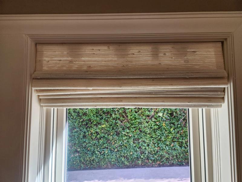 Woven natural fiber Roman shade raised at top of white-trimmed window overlooking hedge