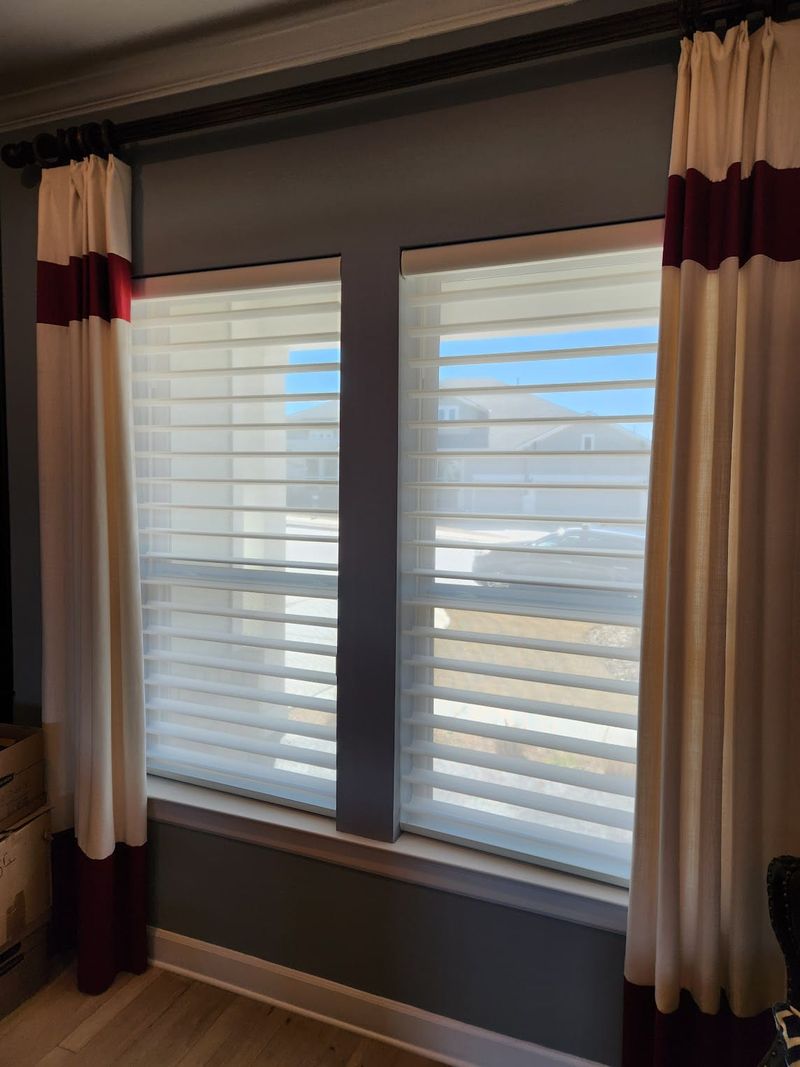 Two white sheer shading blinds on double windows with cream and burgundy drapery panels