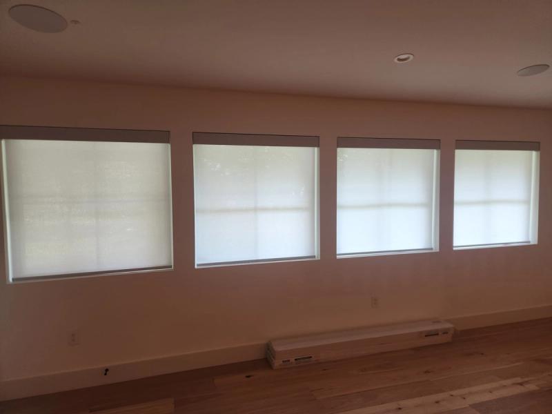 Four white solar roller shades covering square windows in a bright, empty room with hardwood floors