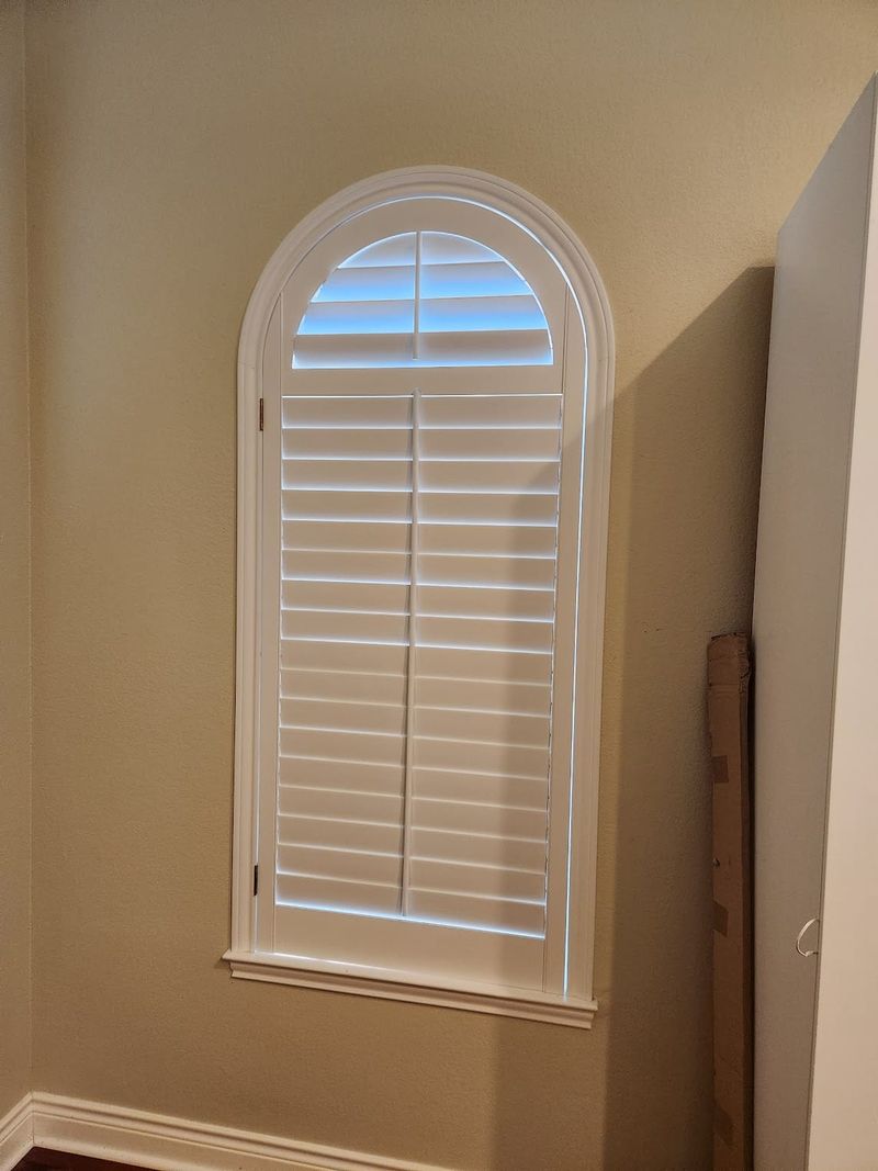 White shutters on a tall arched window in a neutral-toned interior hallway