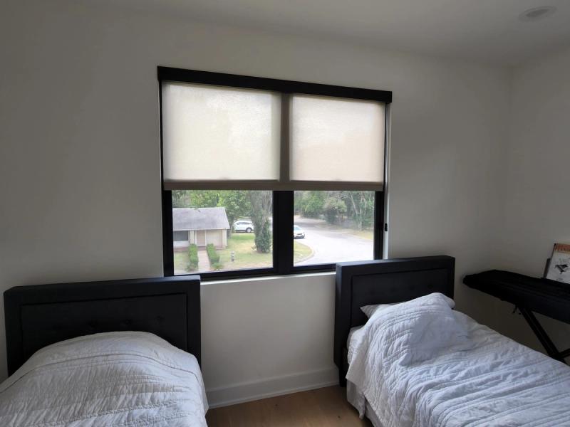 Two beige solar roller shades on wide black-framed window in twin bedroom