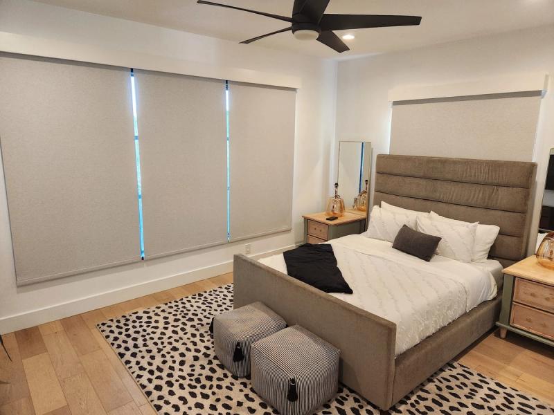 Three light gray roller shades on large windows in a modern bedroom with upholstered bed