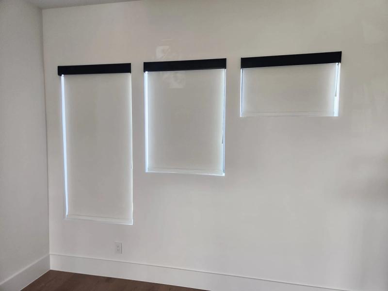 Three white roller shades with black cassettes covering windows of varying sizes in a modern white room