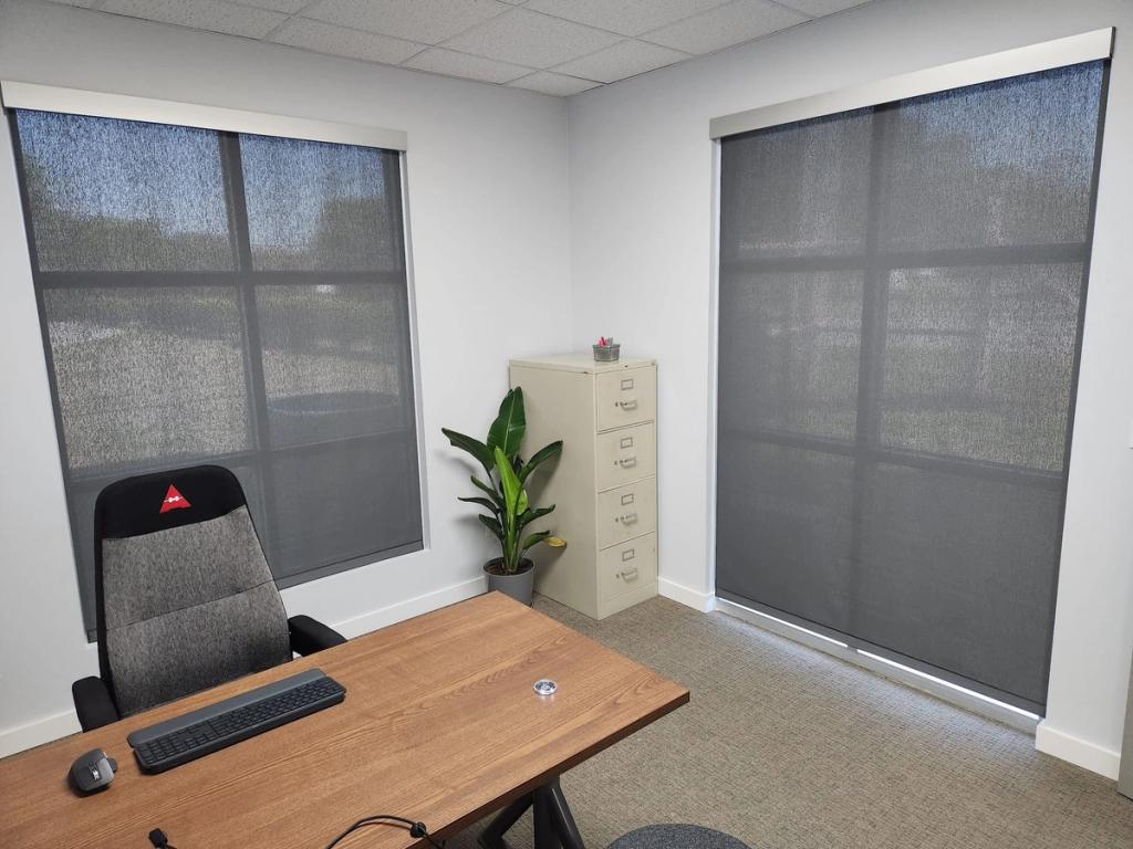 Gray solar roller shades on two large windows in a small office with desk and filing cabinet