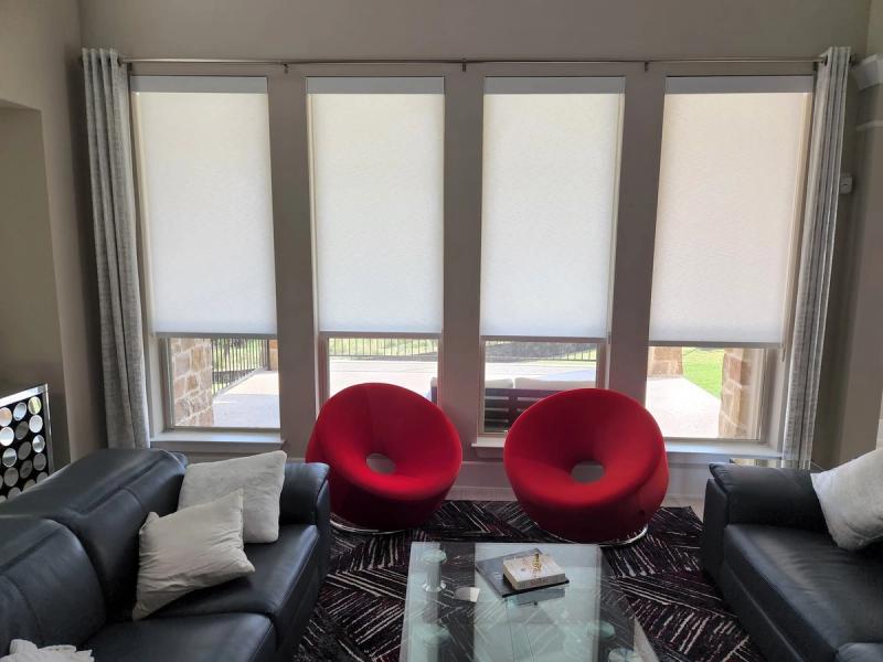 Four white roller shades on large windows in modern living room with red accent chairs