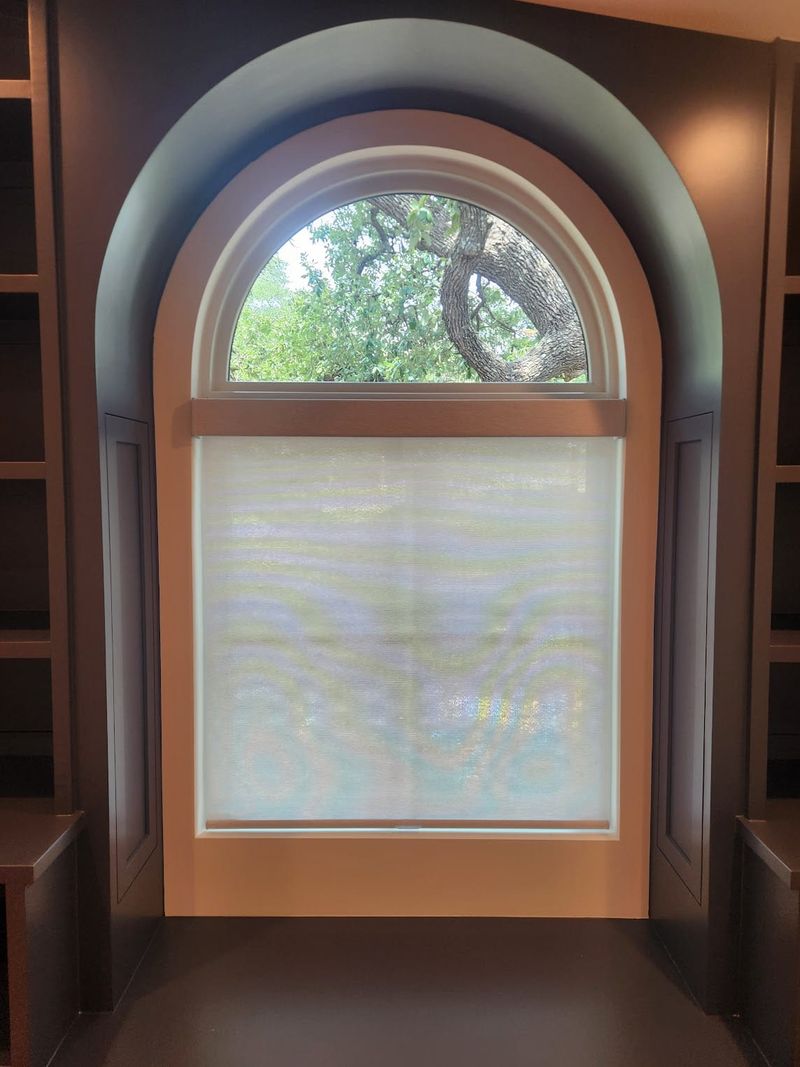 White roller shade on rectangular window below arched transom with tree view