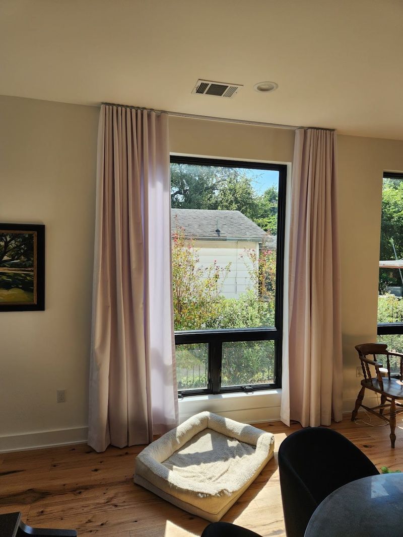 Light pink ripple-fold drapery panels on ceiling-mounted track beside large black-framed window