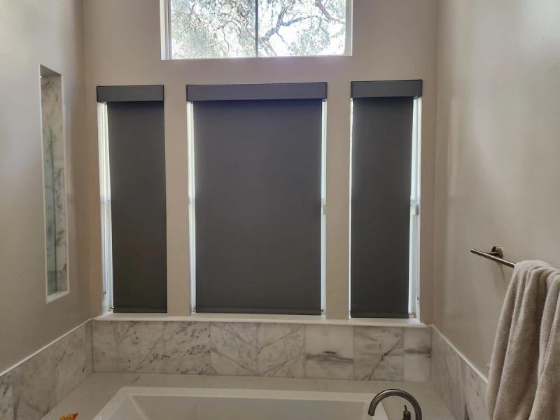 Three dark gray roller shades covering tall windows above a marble bathtub