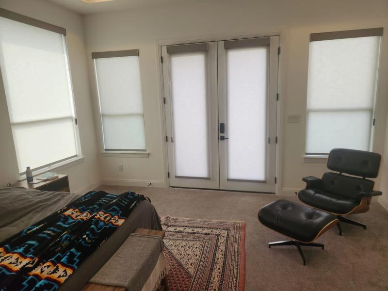 White roller shades on multiple windows and French doors in a modern bedroom with Eames lounge chair