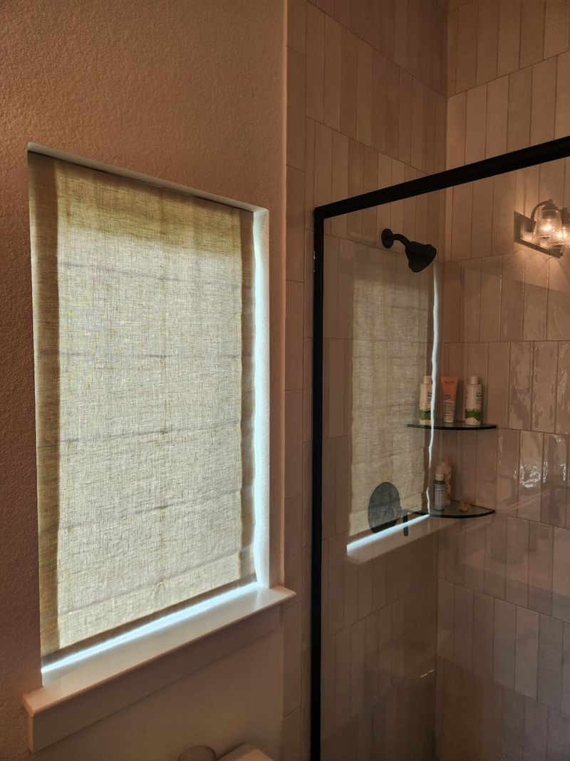 Manual roller shade on bathroom window beside glass shower enclosure with tile walls