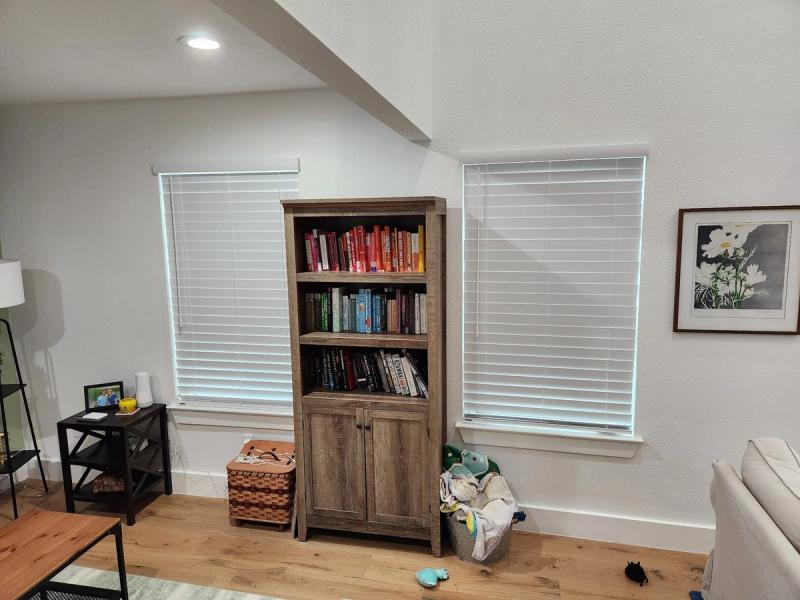 White horizontal blinds on two living room windows flanking a rustic bookcase