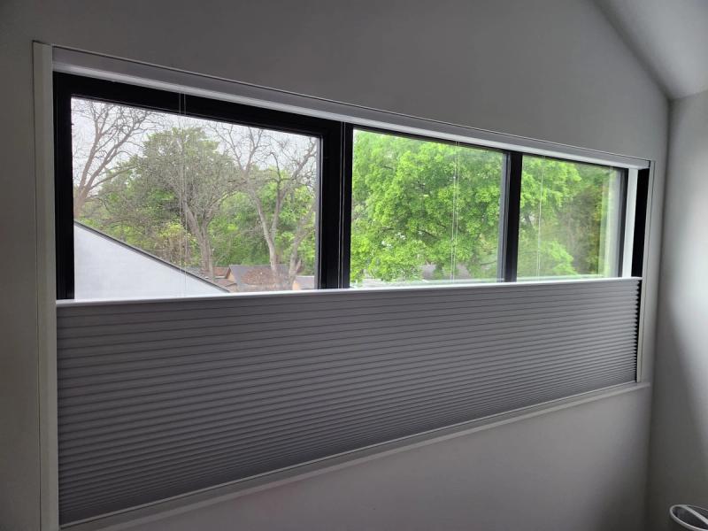 Gray cellular honeycomb shade partially raised on wide horizontal clerestory window overlooking trees