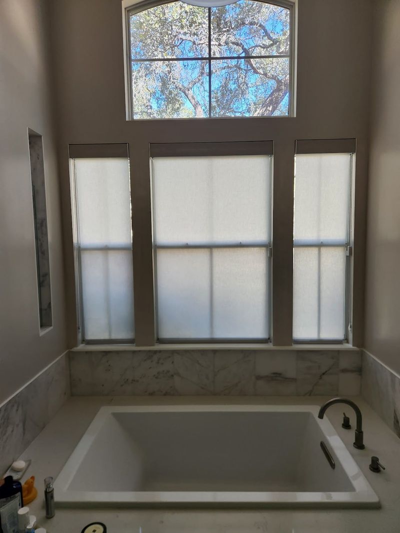 Three white roller shades on tall bathroom windows above marble soaking tub