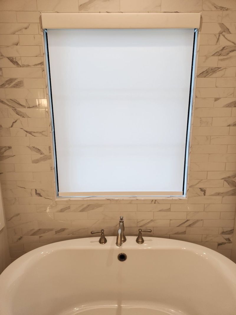 White roller shade fully lowered over bathroom window above soaking tub with marble tile surround