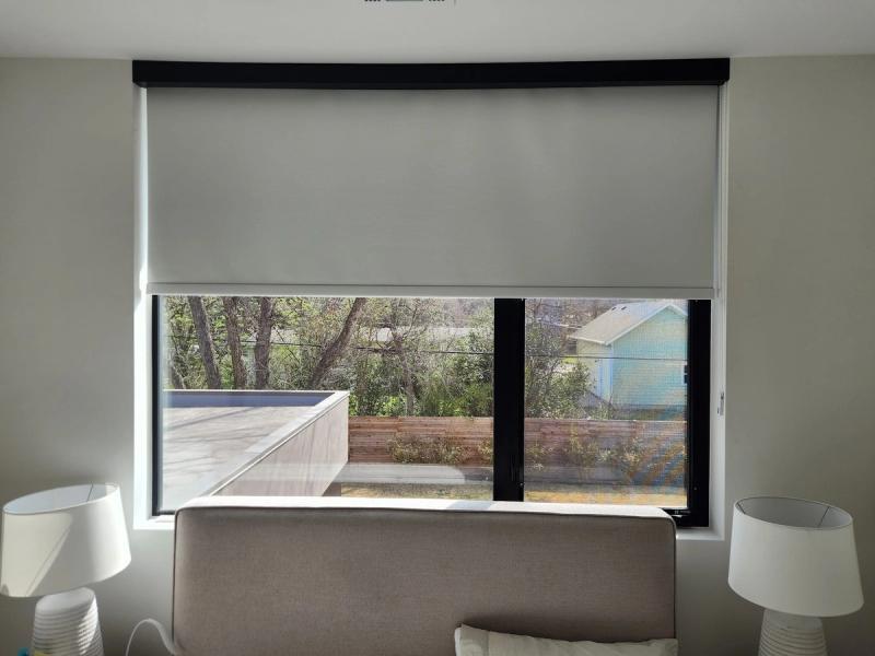 White roller shade partially lowered over large black-framed bedroom window with outdoor patio view