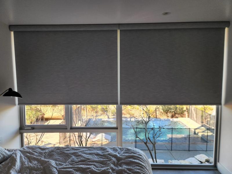 Two gray roller shades partially lowered over large bedroom windows overlooking a pool