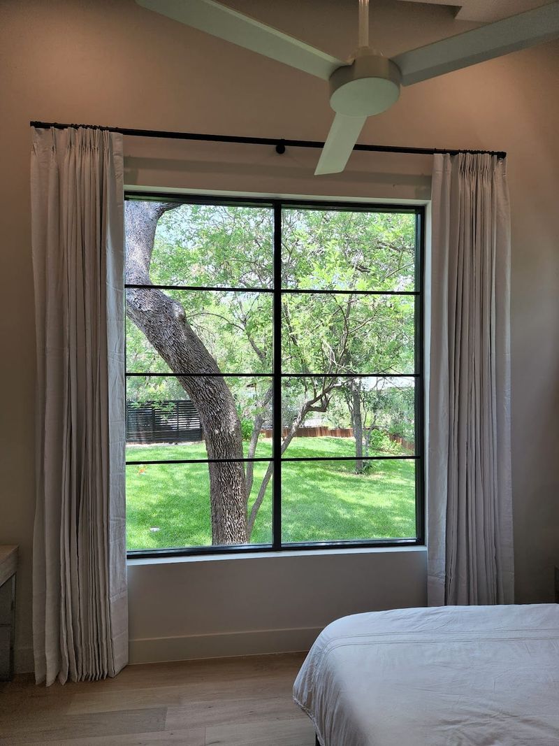 Pinch-pleat white drapery panels on black rod framing large grid window in bedroom