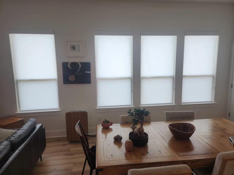 Four white roller shades on bright windows in a modern dining room with wood table