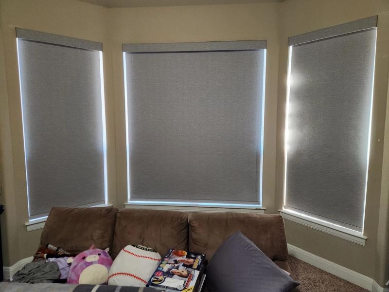 Three gray roller shades on a bay window above a brown sofa with stuffed animals
