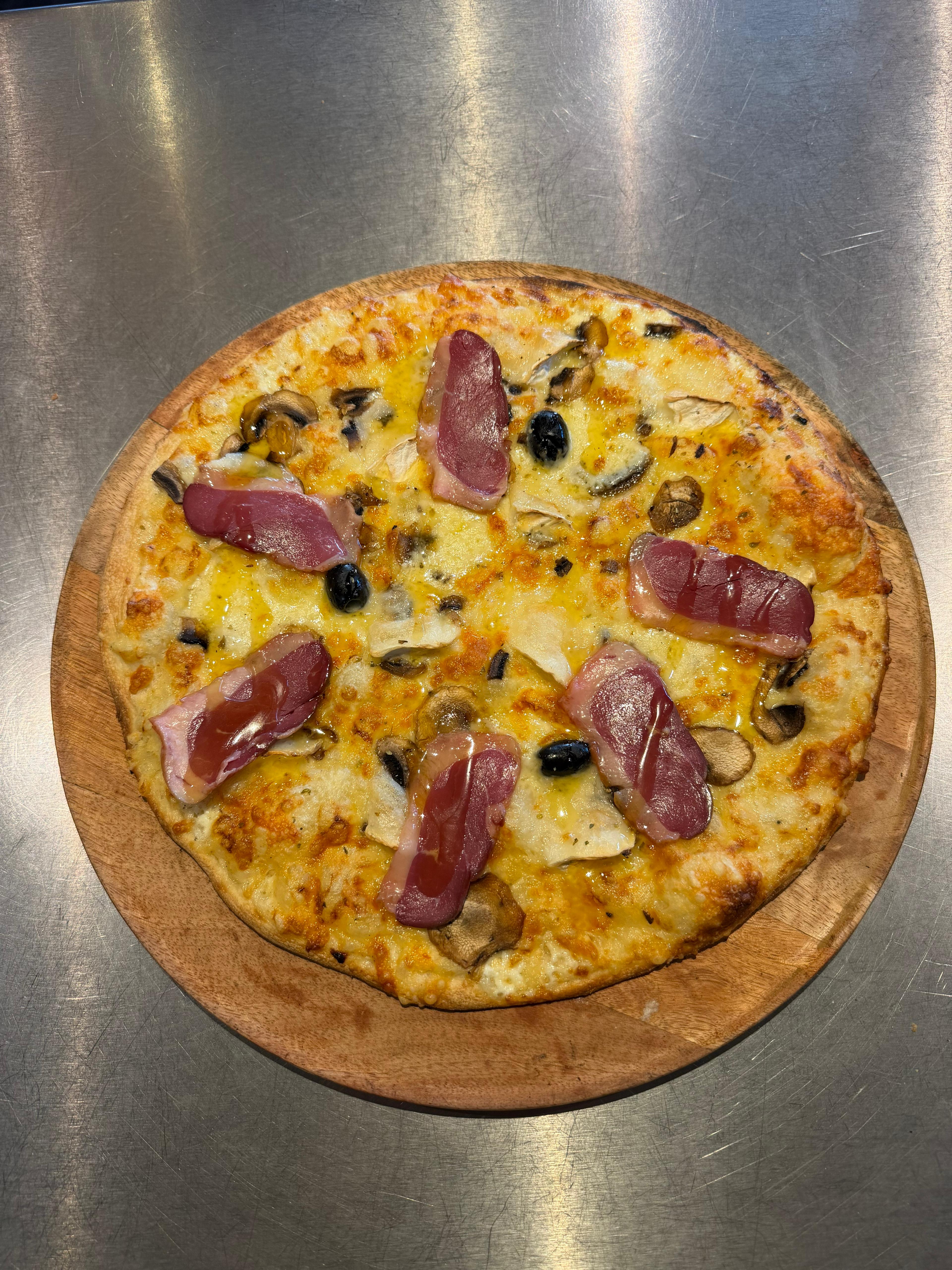 Pizza Duck artisanale garnie de magret de canard, champignons, fromage de chèvre, filet de miel, fromage fondu et olives noires, présentée sur une planche en bois à la pizzeria Chez Matteo au Porge.