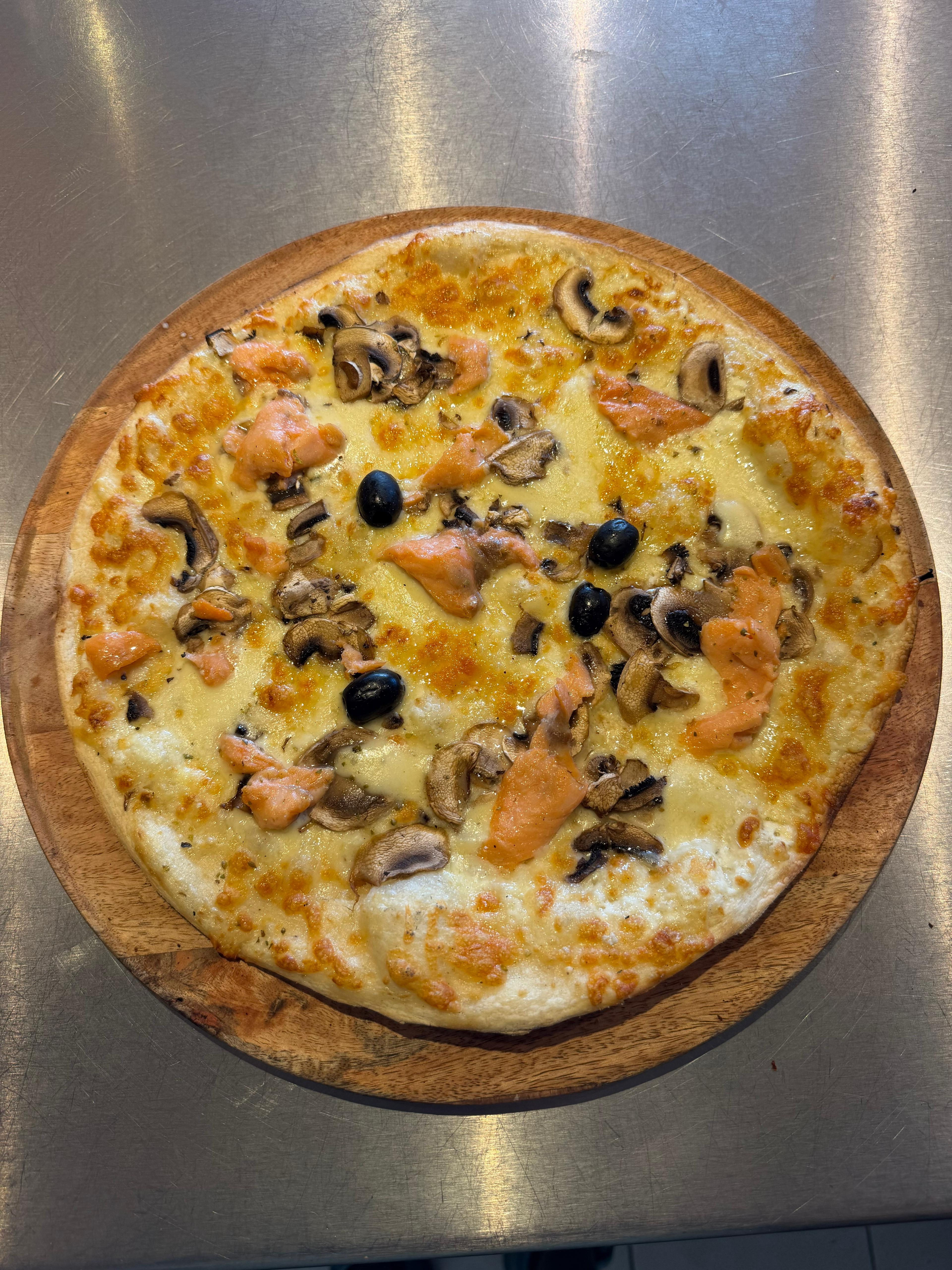 Pizza Norvégienne artisanale garnie de saumon fumé, de champignons, de mozzarella fraîche, d'un mélange d'emmental et d'olives noires, présentée sur une planche en bois à la pizzeria Chez Matteo au Porge.