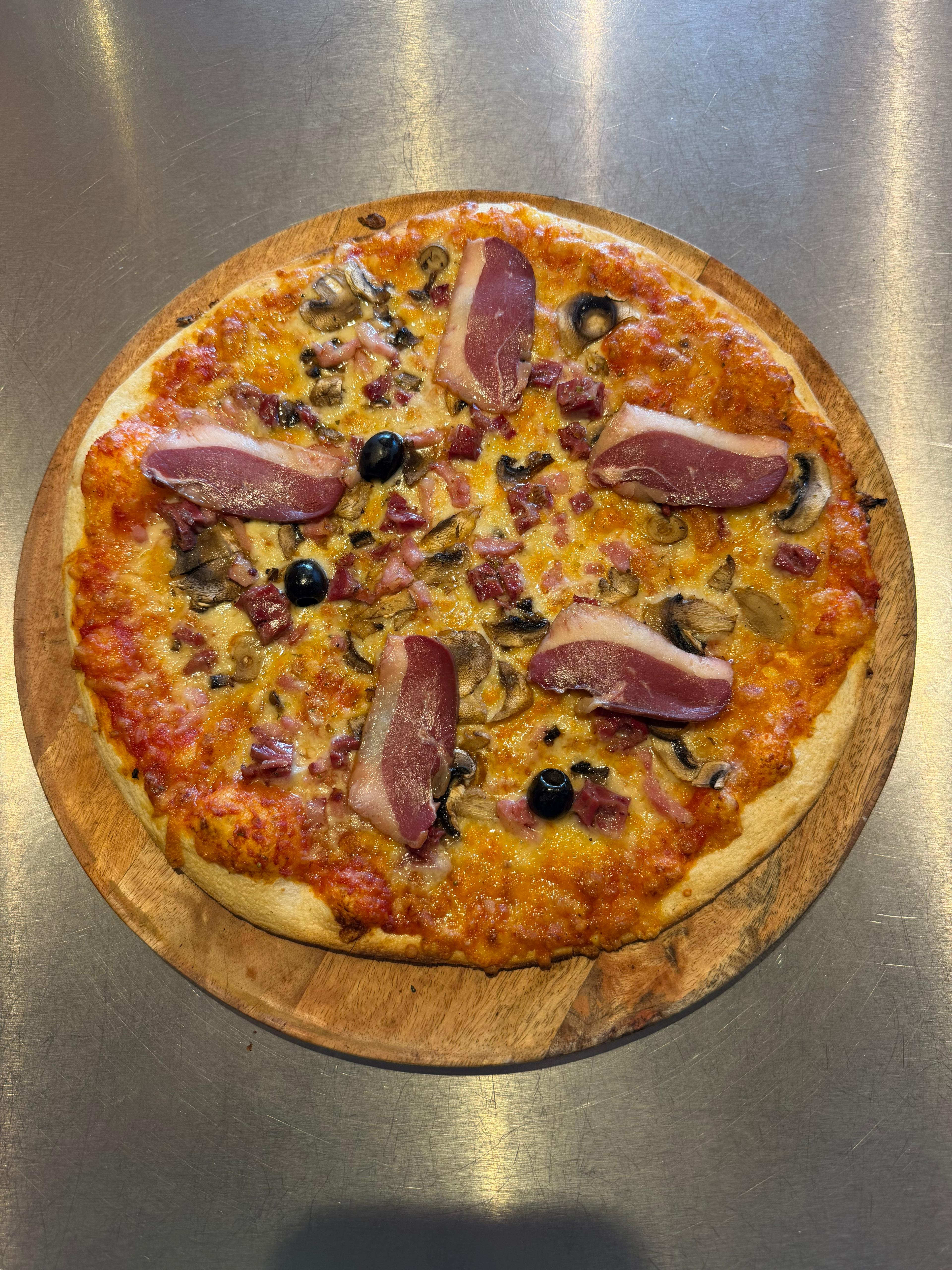 Pizza Landaise artisanale garnie de magret de canard, gésiers, lardons, champignons, fromage fondu et olives noires, présentée sur une planche en bois à la pizzeria Chez Matteo au Porge.