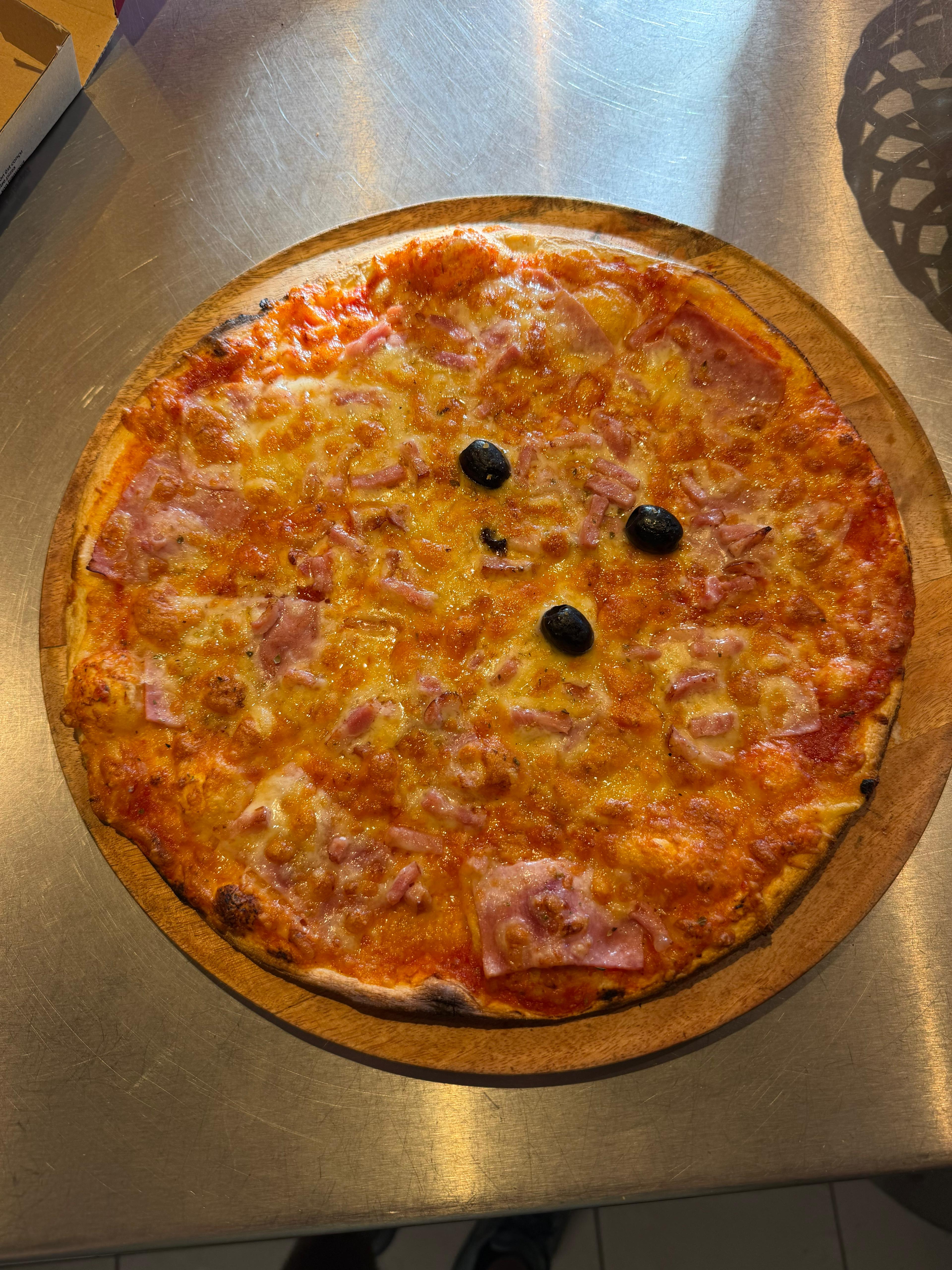 Pizza Reine artisanale classique garnie de jambon blanc, d'un savoureux mélange d'emmental et de mozzarella fondus, et d'olives noires, présentée sur une planche en bois à la pizzeria Chez Matteo au Porge.