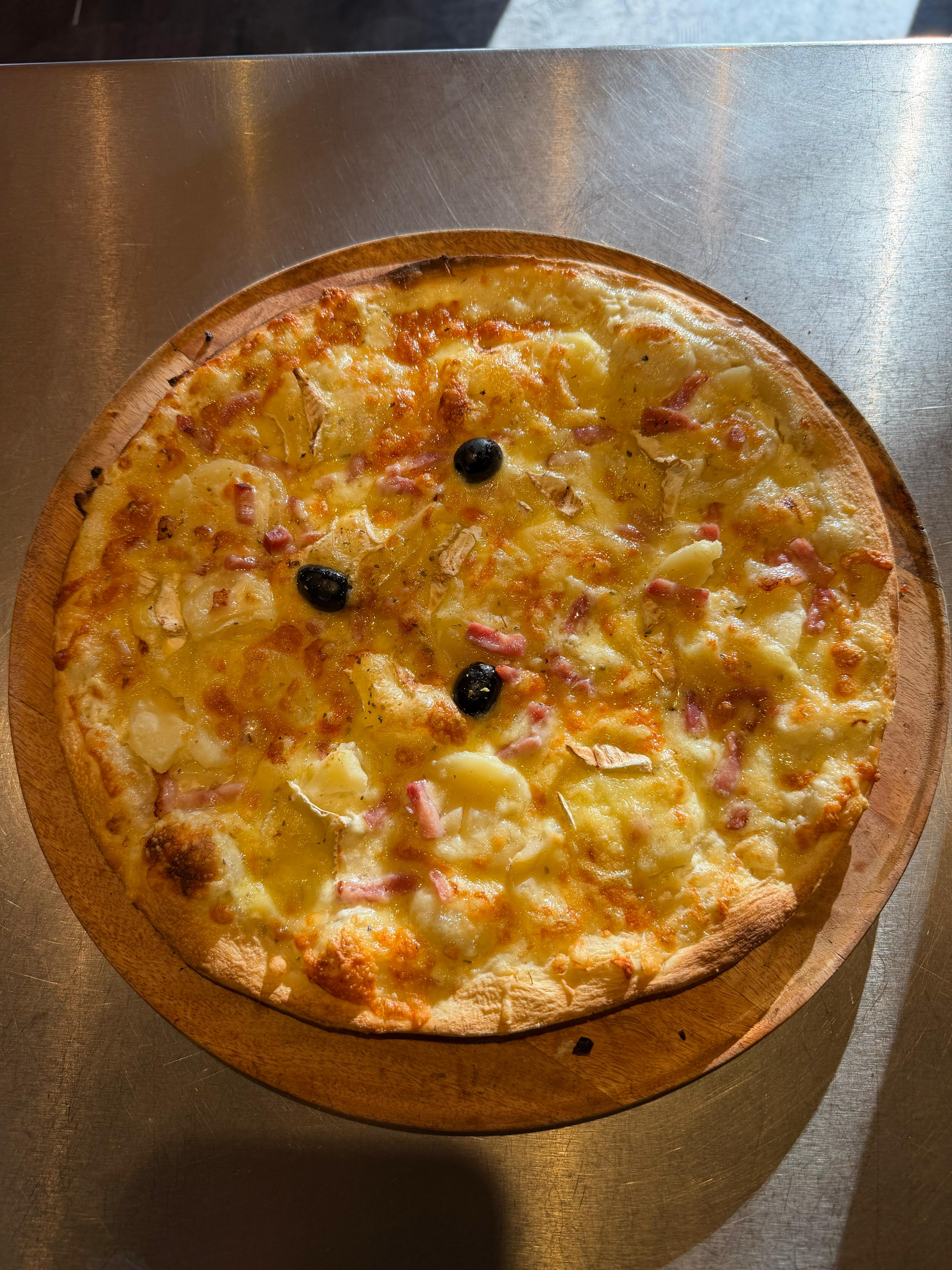 Pizza Normande artisanale garnie de pommes de terre, lardons, camembert, fromage fondu et olives noires, présentée sur une planche en bois à la pizzeria Chez Matteo au Porge.