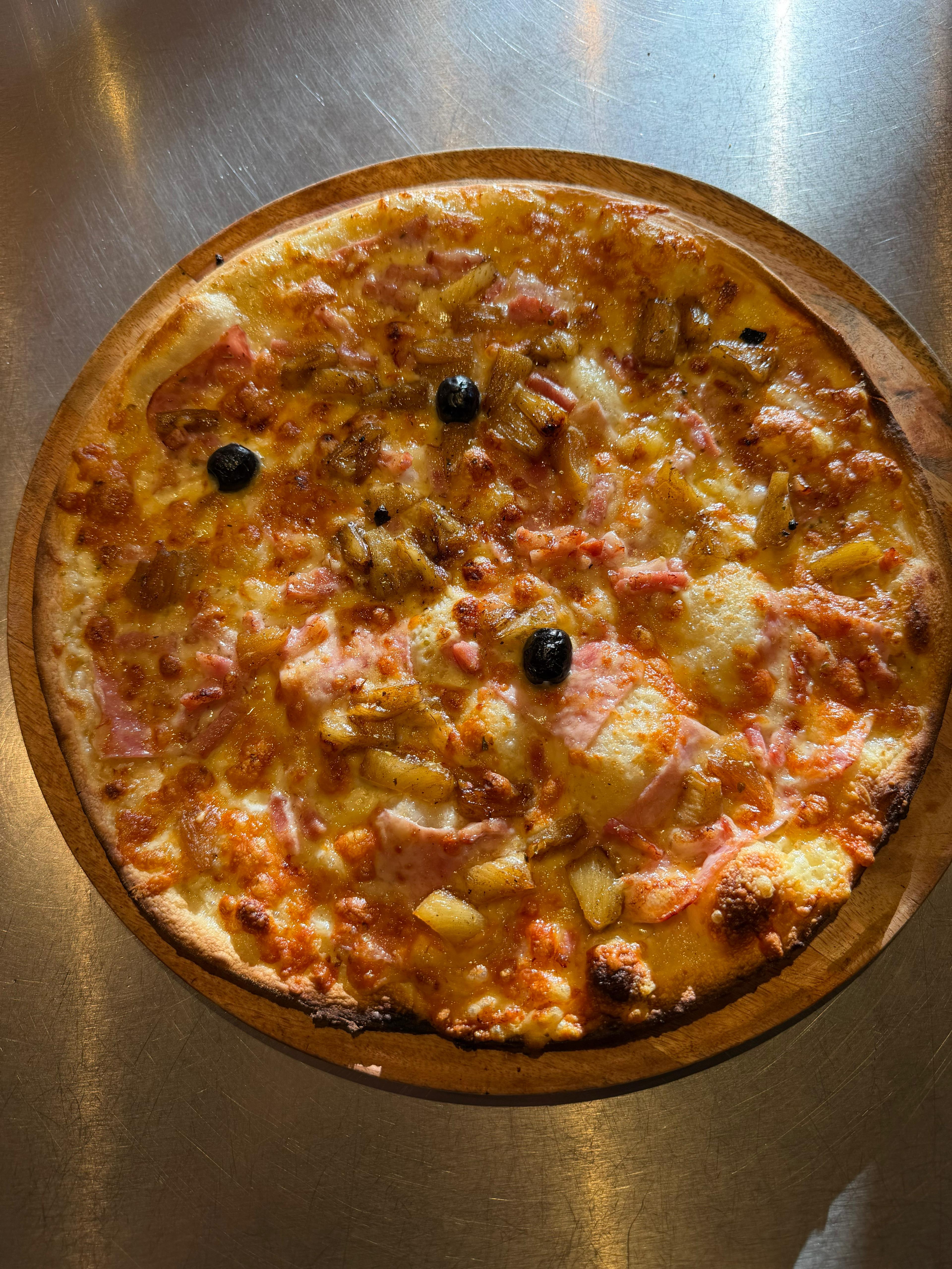 Pizza Hawaïenne artisanale garnie de jambon, de morceaux d'ananas, de fromage fondu et d'olives noires, présentée sur une planche en bois à la pizzeria Chez Matteo au Porge.
