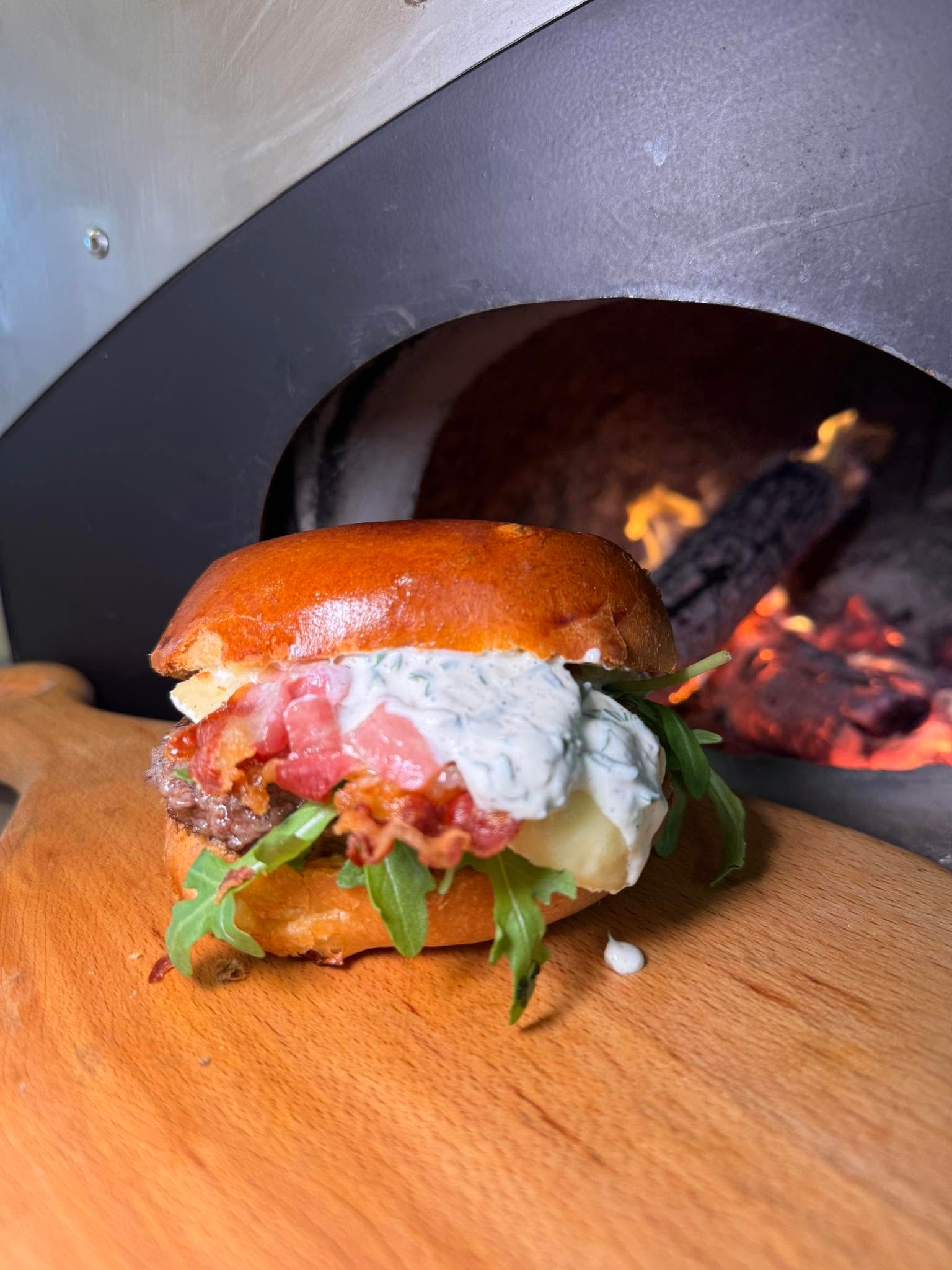 Burger artisanal gourmand avec pain brioché, bacon, roquette et sauce aux herbes, présenté sur une planche en bois devant les flammes du four traditionnel de Chez Matteo au Porge.