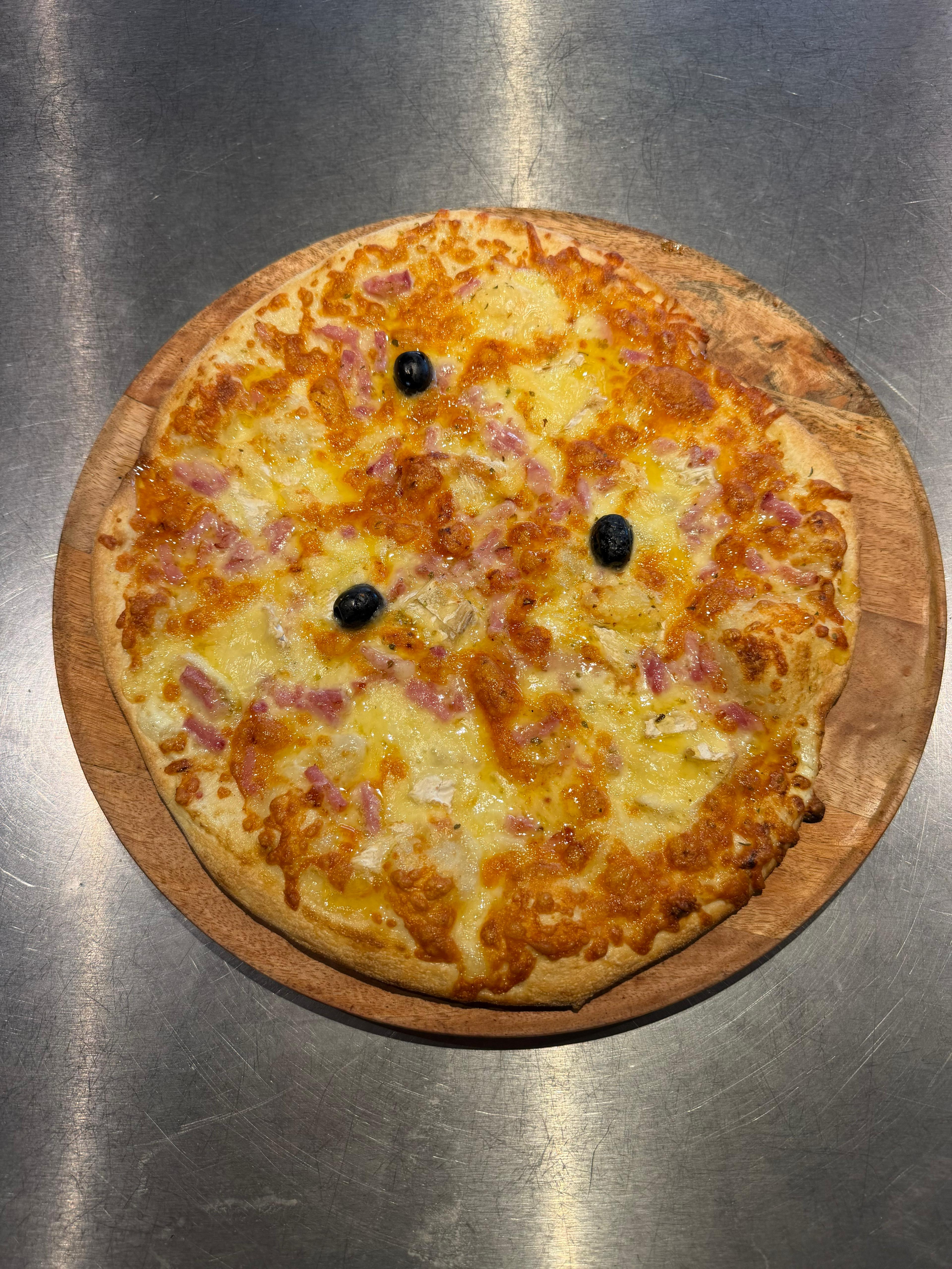 Pizza Normande artisanale garnie de lardons, morceaux de camembert, fromage fondu et olives noires, présentée sur une planche en bois à la pizzeria Chez Matteo au Porge.