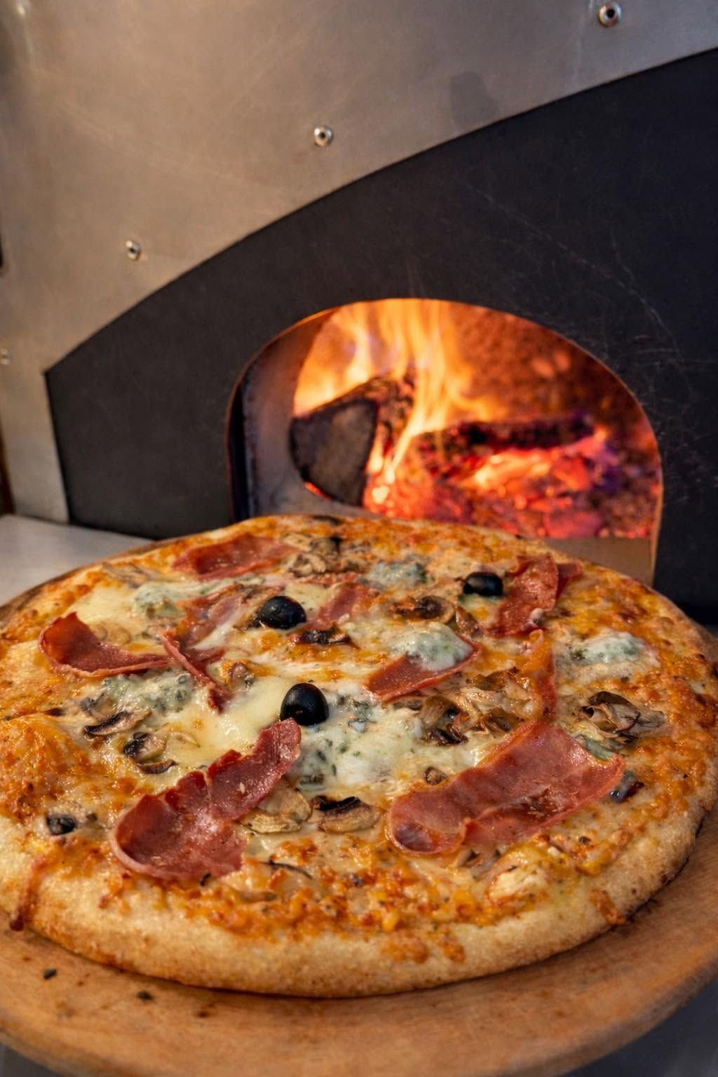 Pizza artisanale à la croûte dorée garnie de jambon, champignons, olives noires et fromage fondu, présentée sur une pelle devant un four à bois traditionnel.