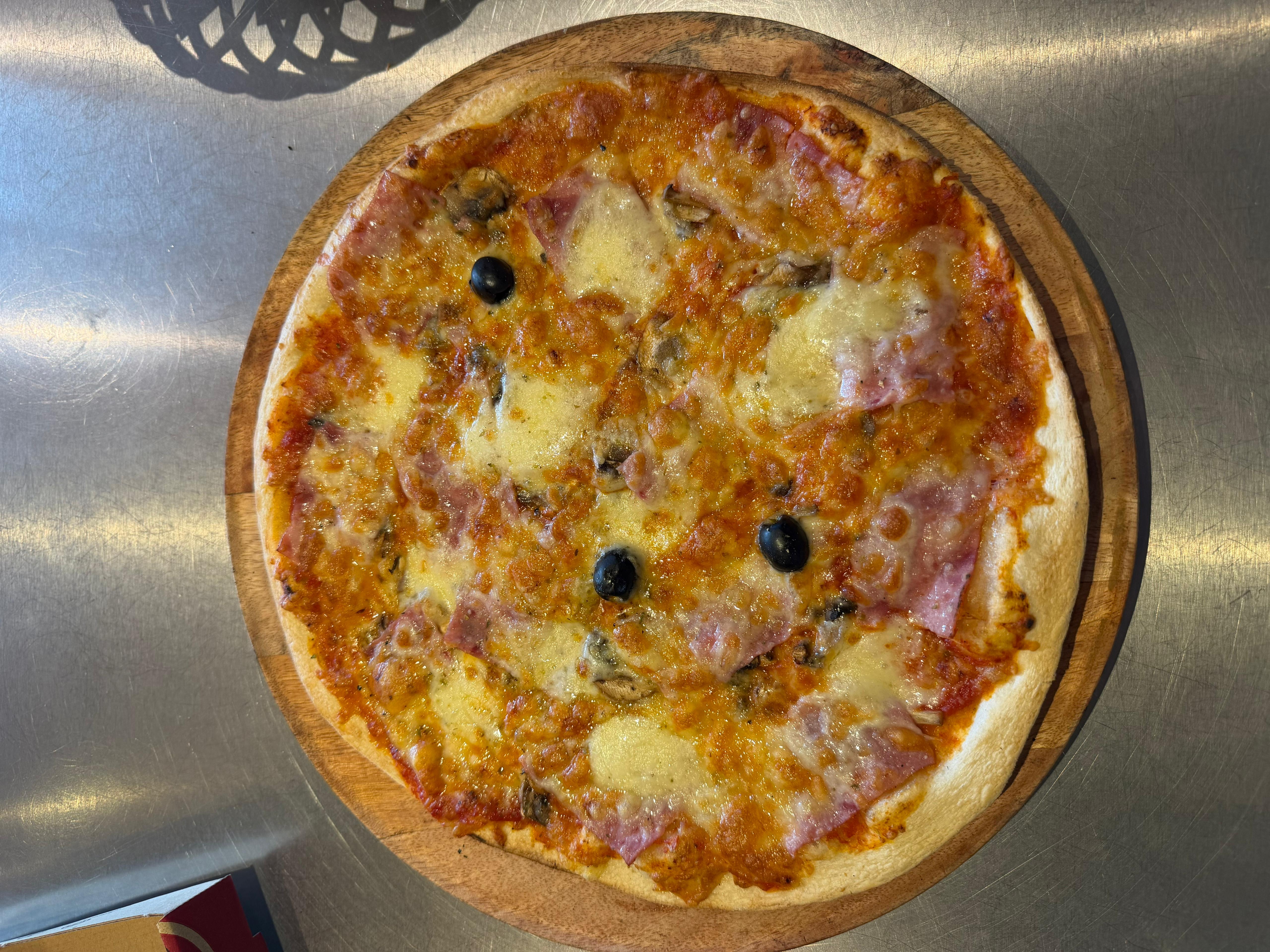 Pizza Bambino artisanale garnie de jambon blanc, champignons, mélange de fromages fondus et olives noires, présentée sur une planche en bois à la pizzeria Chez Matteo au Porge.