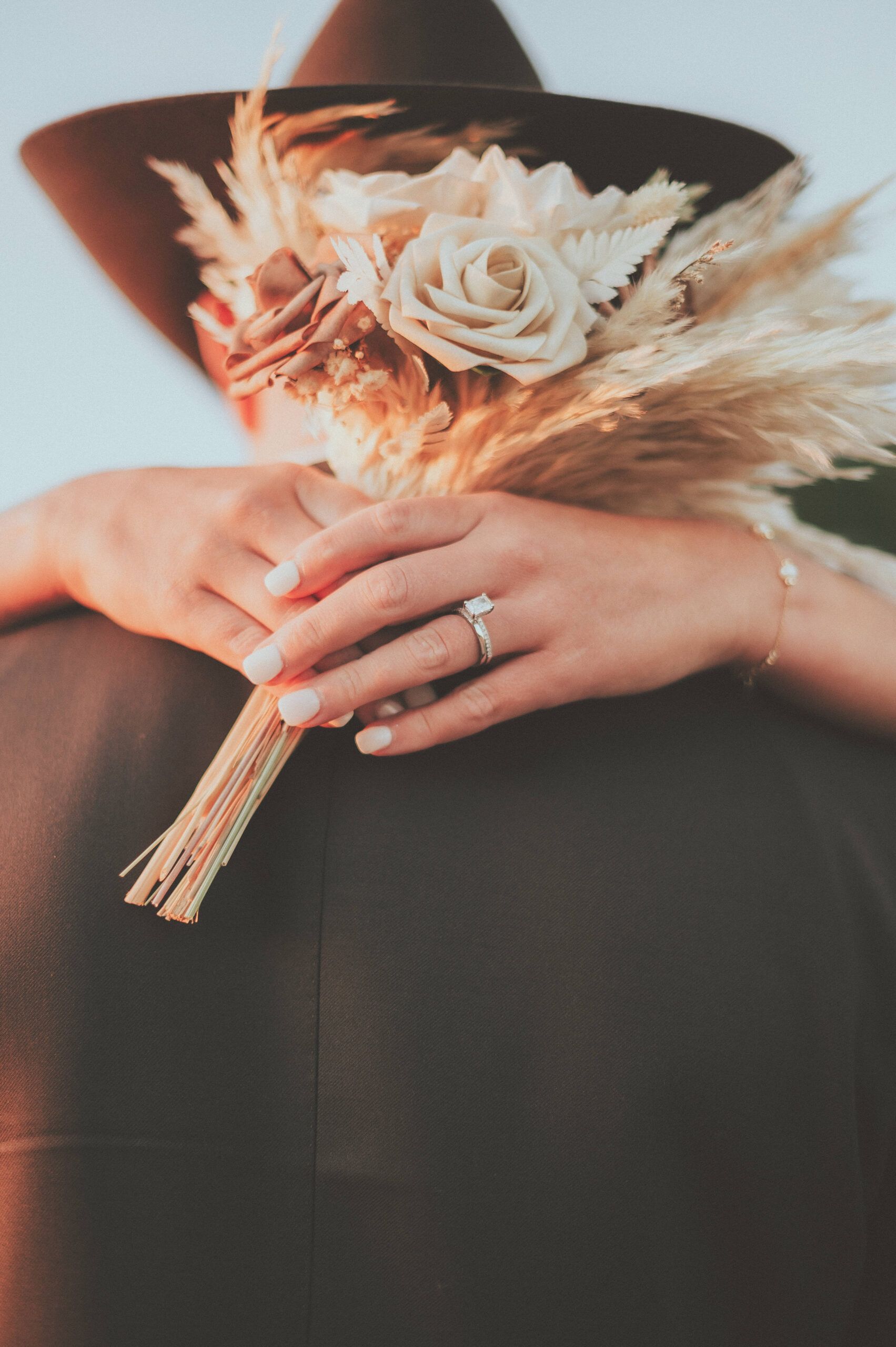 A close-up of a ring, hands, and a peach-and-cream bouquet against a dark suit.