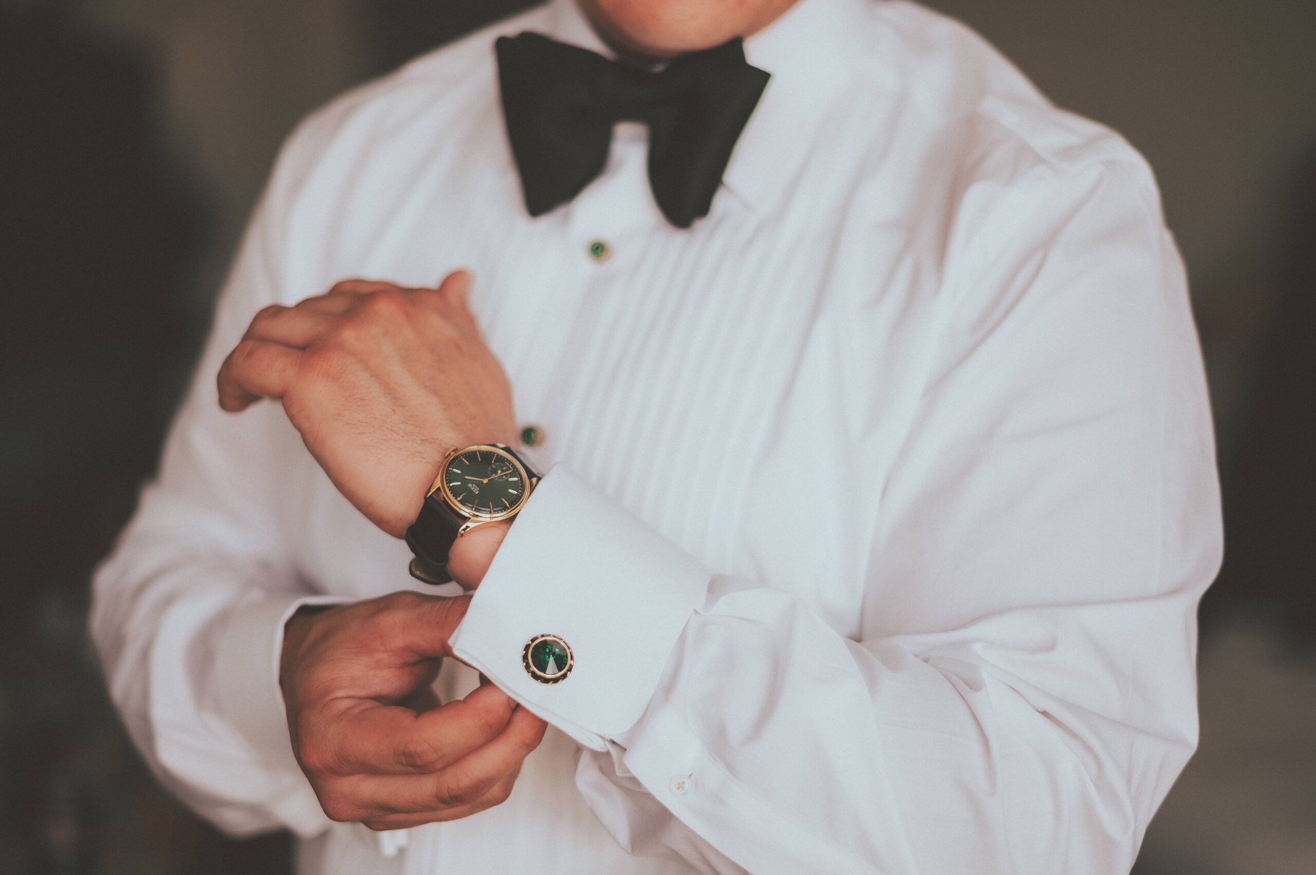 A close-up of a shirt cuff, wristwatch, and bow tie during wedding preparations.