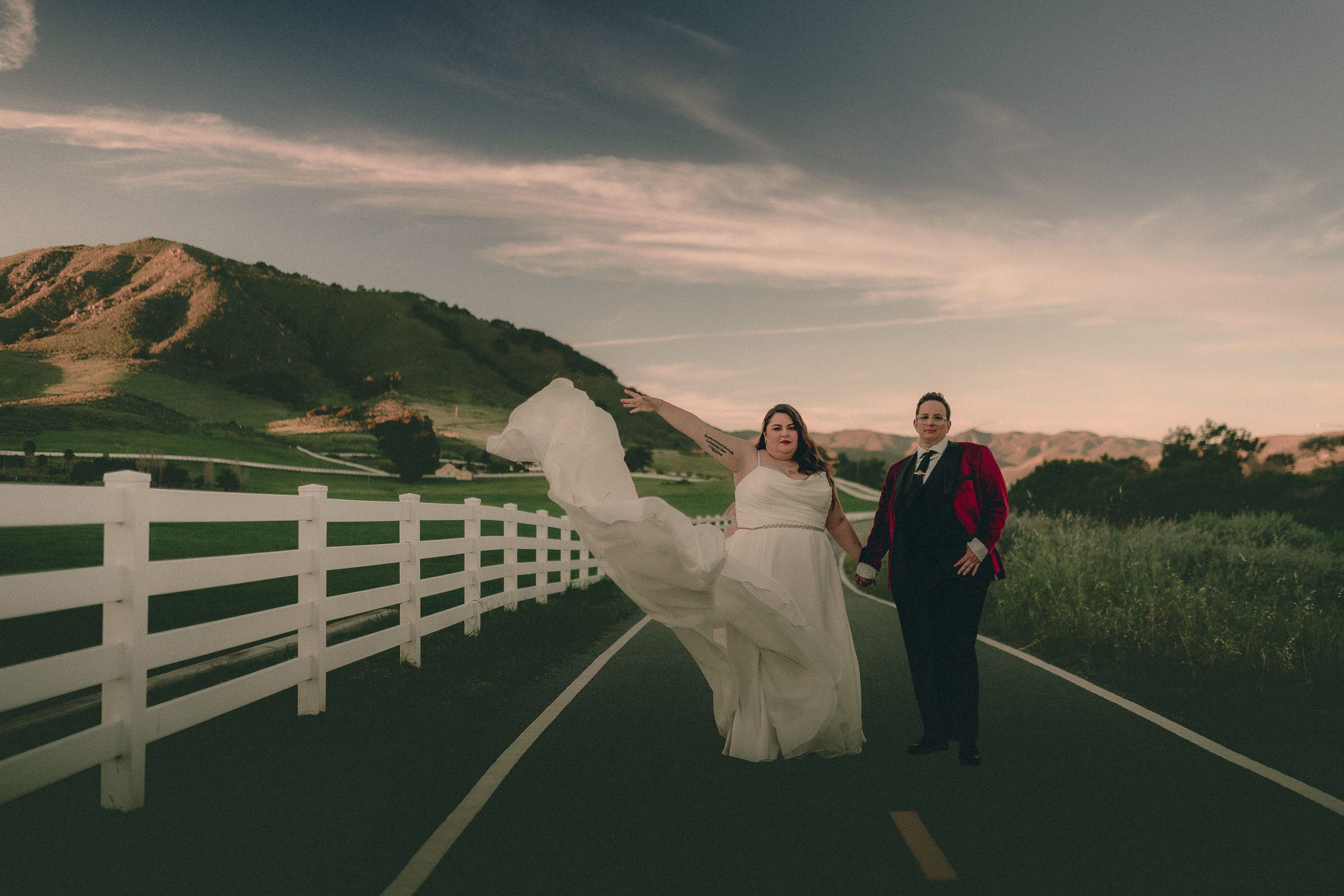A couple hold hands on a country road at sunset, with a flowing wedding dress, burgundy jacket, white fence and rolling hills.