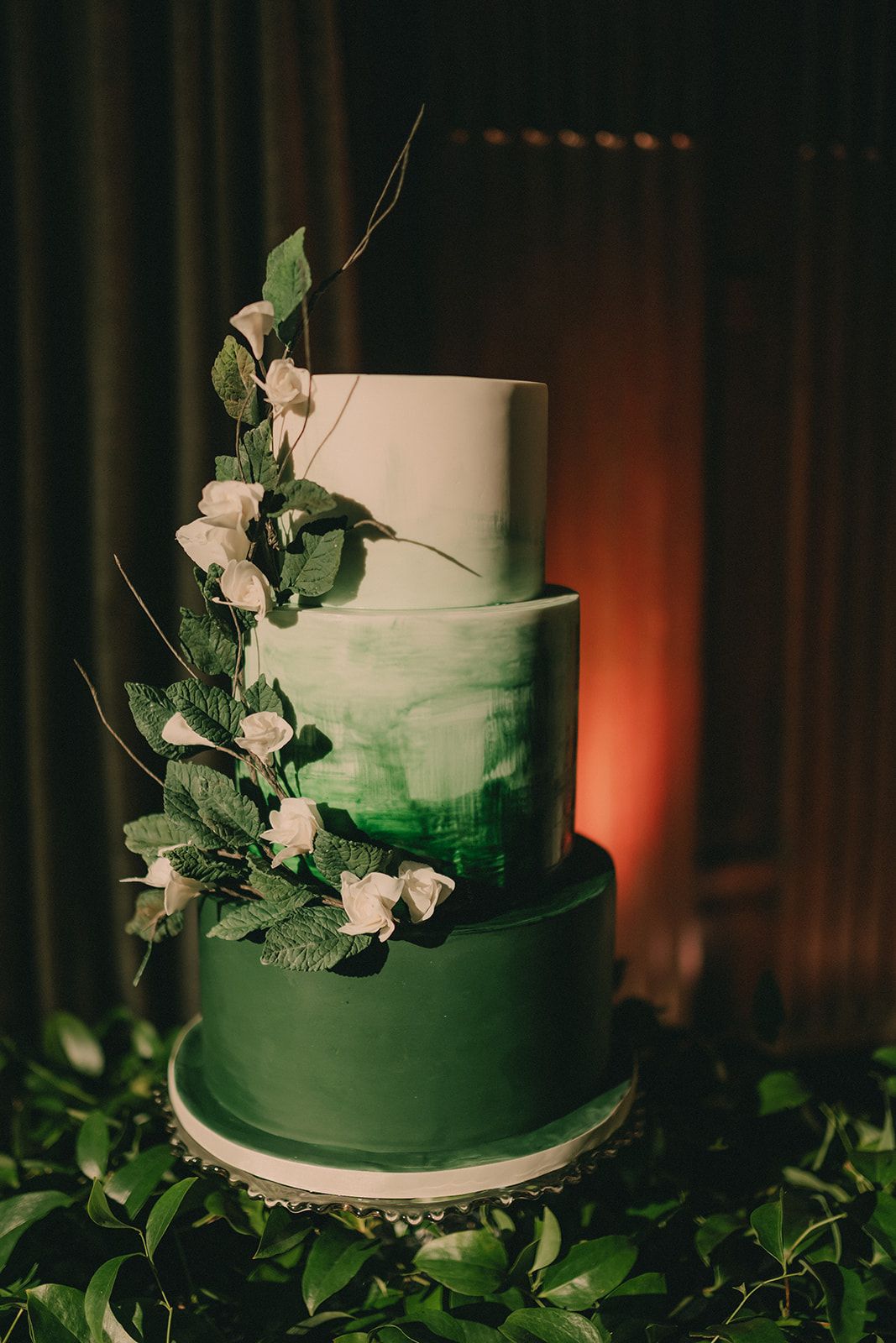 Three-tier green wedding cake decorated with white flowers and leafy vines.