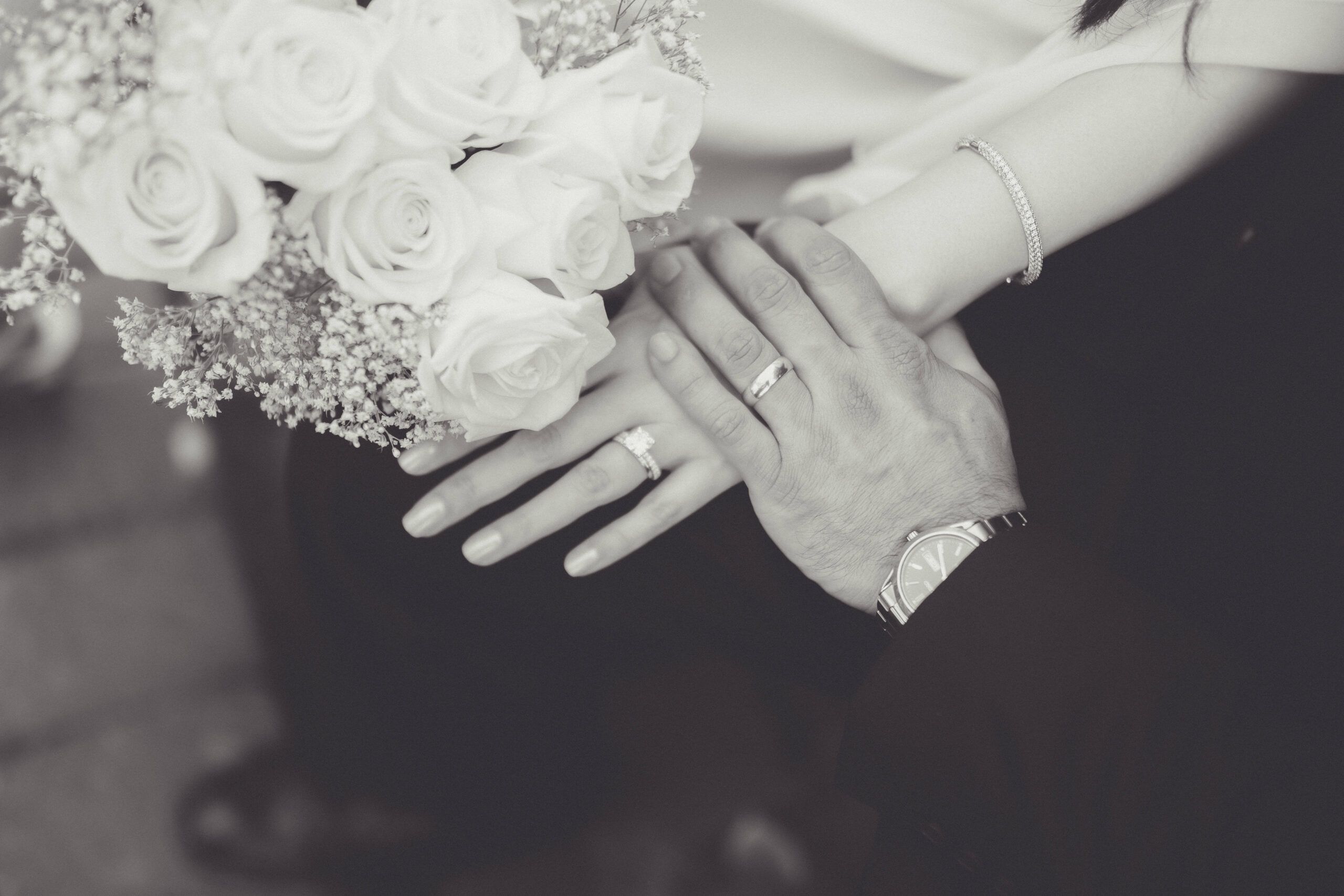 Black-and-white close-up of joined hands, wedding rings, and a bouquet of white roses.