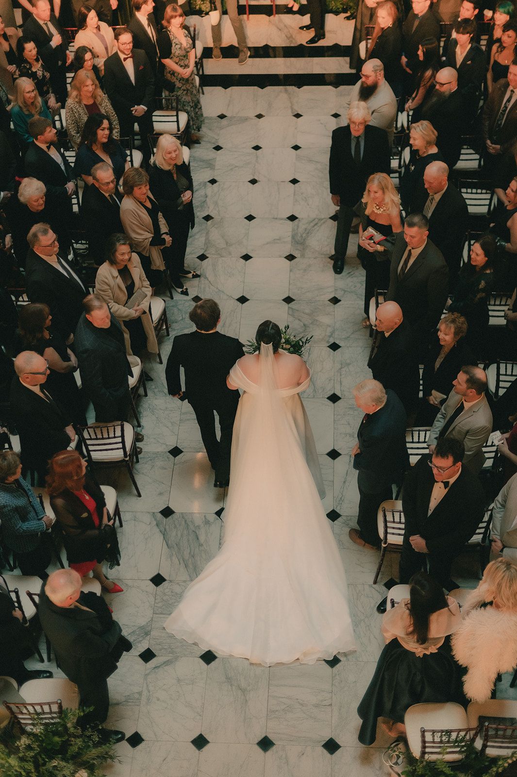 Overhead view of Anne walking into the marble ceremony aisle as guests look on.