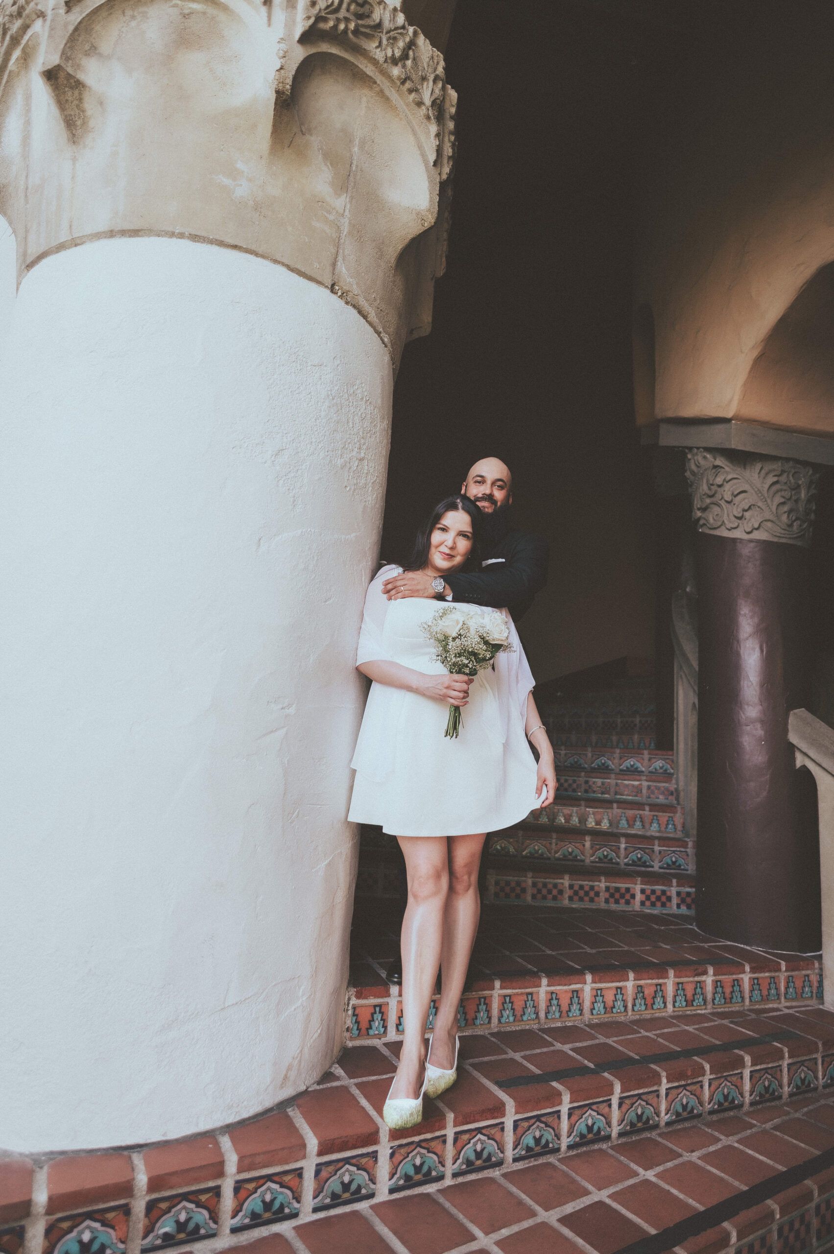 Newlyweds embracing on tiled steps beneath carved stone columns.