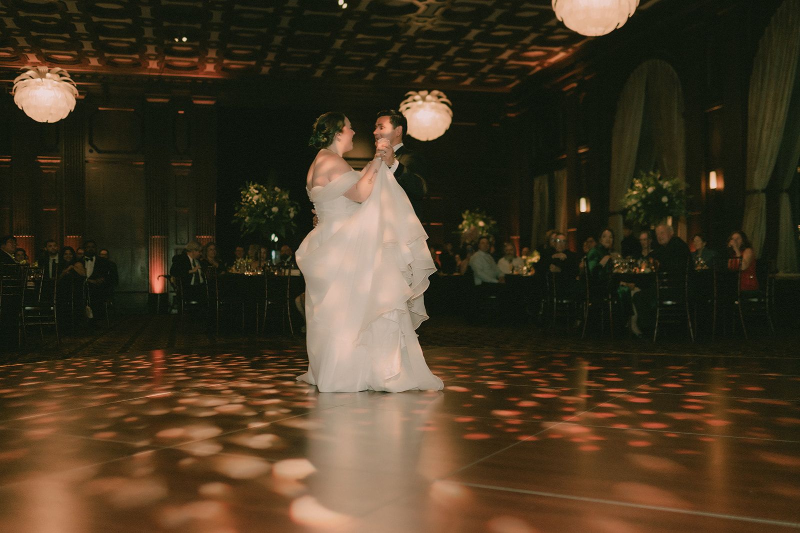 Anne and Charles laugh together during their first dance beneath patterned amber light.