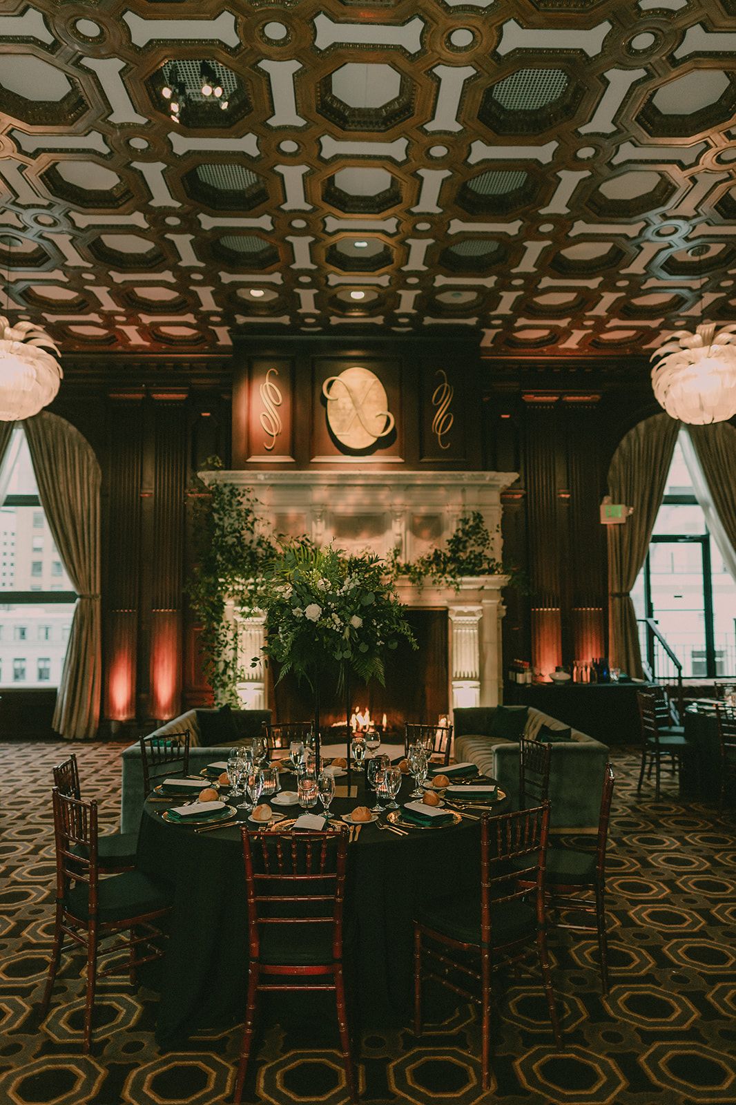 Candlelit reception table beside a marble fireplace beneath an ornate geometric ceiling.