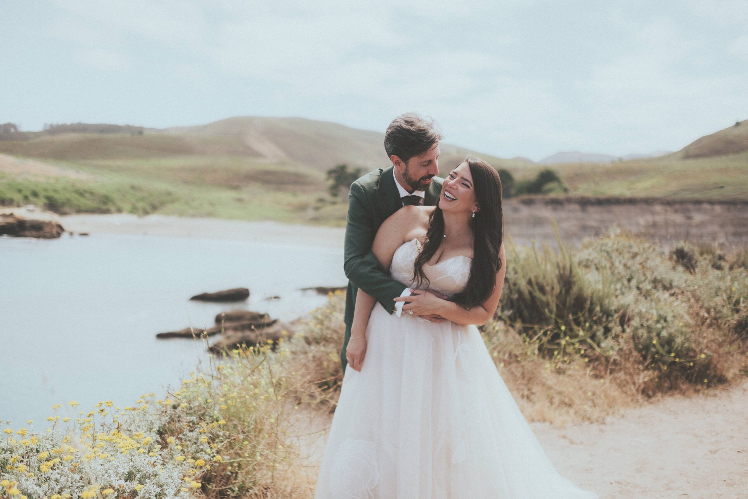 Newlyweds laughing together beside the water with soft green hills behind them.