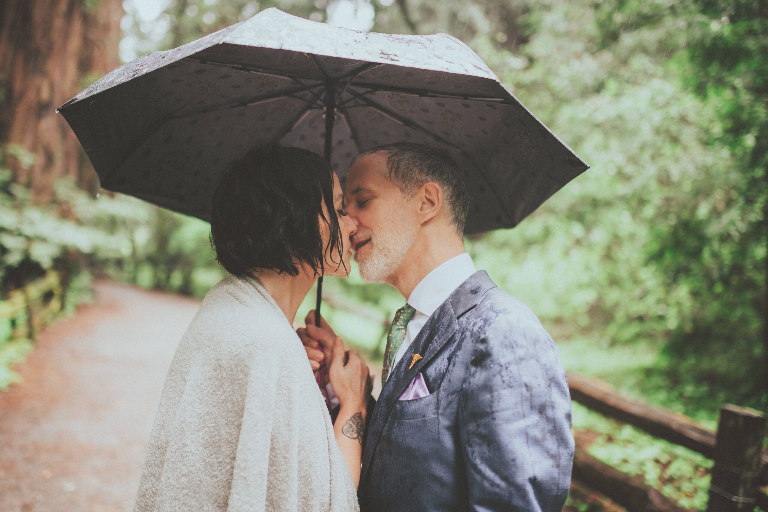 A couple sharing a kiss under an umbrella on a rainy woodland path.