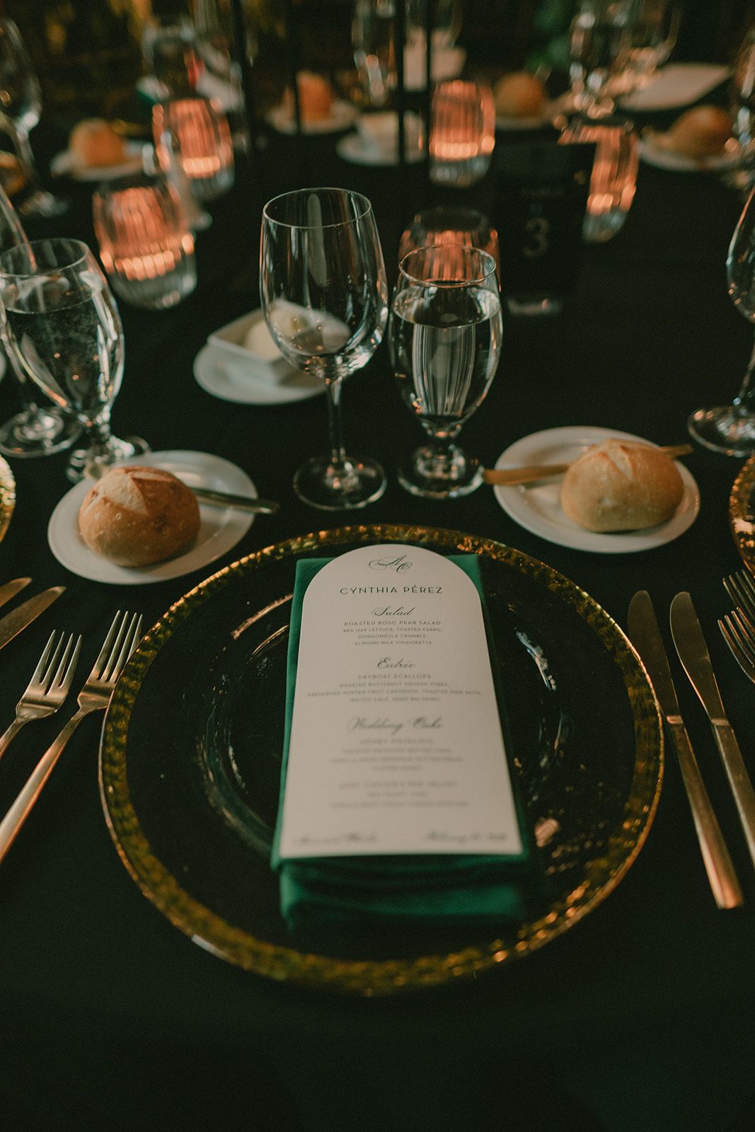 Candlelit place setting with a deep-green napkin, dark glass charger and wedding menu.