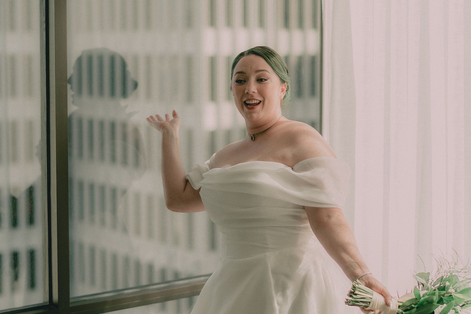 Anne laughs beside a hotel window while holding a loose green-and-white bouquet.